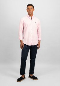 Camicia a maniche lunghe rosa chiaro con taschino sul petto, abbinata a pantaloni blu scuro e scarpe slip-on nere con dettagli intrecciati.