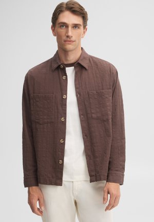 Mann trägt ein braunes, strukturiertes Hemd mit Knöpfen über einem weißen T-Shirt und hellen Hosen vor einem schlichten hellen Hintergrund.