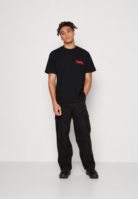 Schwarzes T-Shirt mit roter Grafik auf der Brust, kombiniert mit schwarzen Cargo-Hosen. Kurze Ärmel, runder Ausschnitt und lässige Passform.