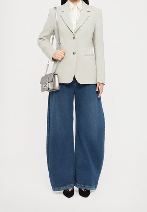 Donna che indossa un blazer bianco, una camicia a righe, jeans blu a gamba larga, scarpe nere con punta e porta una piccola borsa a tracolla argentata.