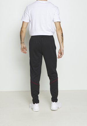 Person steht mit dem Rücken zur Kamera, trägt ein weißes T-Shirt, schwarze Hose mit rotem Nahtdetail und weiße Sneakers vor einfarbigem Hintergrund.