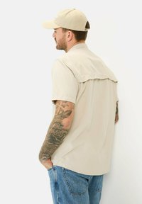 Hellbeige Kurzarmhemd mit einer Aussparung auf der Rückseite, kombiniert mit Jeans. Das Model trägt eine passende beige Kappe und hat sichtbare Tattoos.