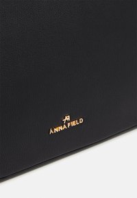 Borsa in pelle nera testurizzata con logo dorato "ANNA FIELD". Forma rettangolare compatta con bordi lisci e senza hardware visibile.