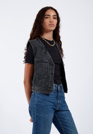 Gilets sans manches en denim noir au design ajusté, avec des poches avant. Associé à une chemise noire et un jean bleu taille haute.