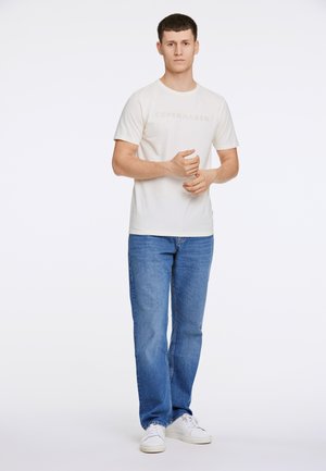 Hellbeige T-Shirt mit "KOPENHAGEN" in einem etwas dunkleren Farbton bedruckt, kombiniert mit blauen Jeans und weißen Turnschuhen.