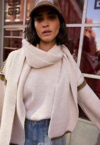 Beige Strickpullover mit einem Schalkragen und offener Vorderseite, ausgestattet mit einem umgeschlagenen Saum und betonten Ärmeln, kombiniert mit hoch taillierten Jeansshorts.