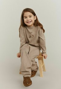 Bruine gebreide trui met kersontwerp, bijpassende bruine gebreide broek en bruin suède laarzen met strassaccenten, zittend op een houten kruk.