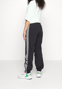 Černé sportovní kalhoty s bílými pruhy po stranách, elastickým pásem a logem Adidas poblíž kotníku. Kombinované s barevnými sportovními botami.