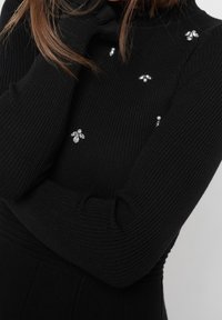 Kvinna som bär en svart ribbad tröja med prydnader i form av juvelerade bin och med händerna delvis över ansiktet.