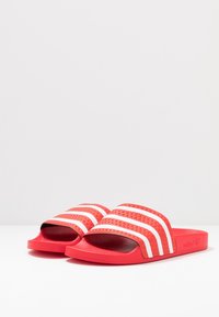 Röda sandaler i gummi med struktur och tre vita ränder över toppen. Bred fotbädd och öppen tådesign för komfort.