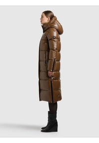 Moteris vilkinti ilgą rudą pūstinį paltą su gobtuvu, šoninėmis užtrauktukais, juodas pėdkelnes ir juodus platforminius aulinukus, stovinti profilio pozicijoje prieš paprastą foną.