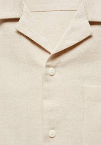 Close-up van een beige gestructureerde stoffen shirt met een notch-kraag, twee witte knopen en een rechthoekige borstzak aan de rechterkant.