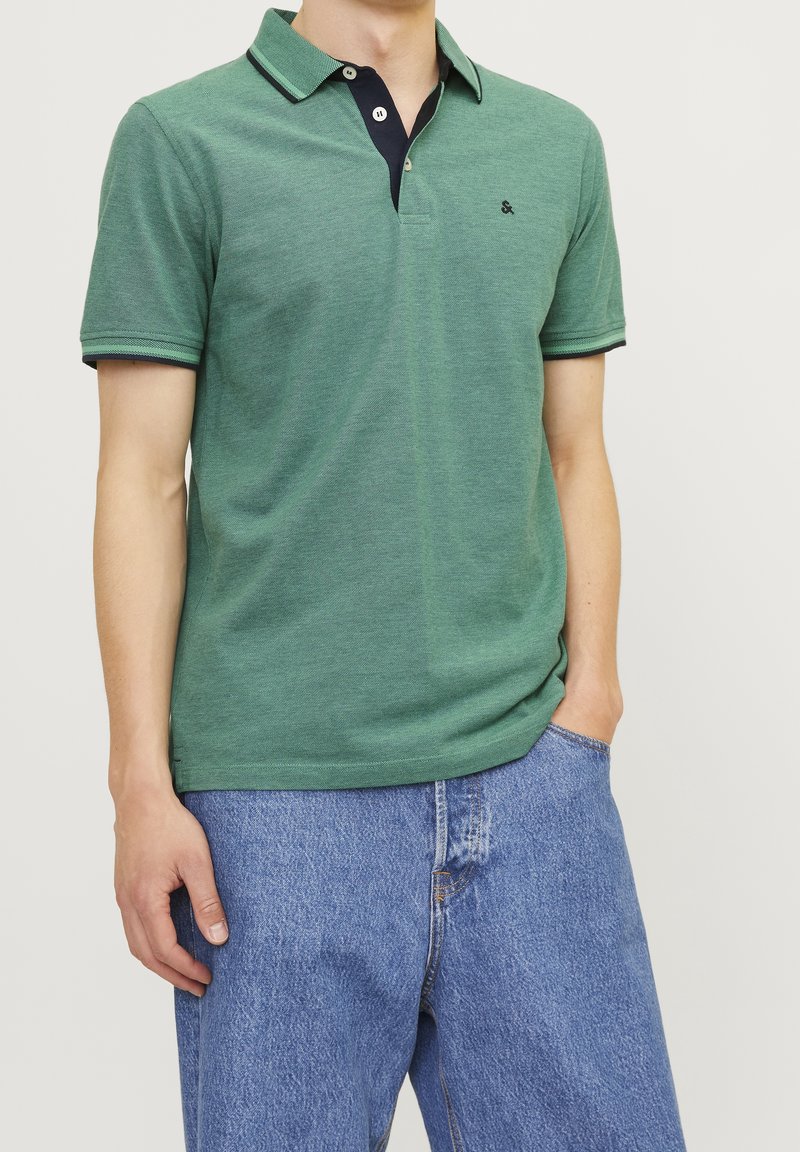Homme portant un polo vert à manches courtes avec un col foncé et un jean bleu, une main dans la poche devant un fond clair uni.