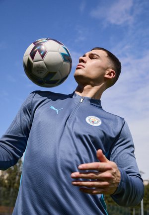 Maglione zip in blu chiaro della Puma con il logo del Manchester City, tessuto testurizzato e motivo sottile, con un pallone da calcio sopra la testa del portatore.