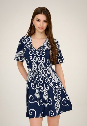 Mladá žena s dlhými hnedými vlasmi, ktorá má na sebe modro-bielo vzorované krátke šaty s výstrihom do V a opaskom v páse.