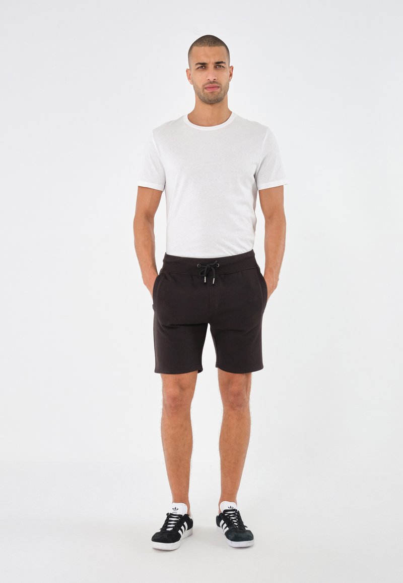 T-shirt bianca a maniche corte e pantaloni corti neri con cordoncino, dal taglio rilassato, abbinati a scarpe da ginnastica nere e bianche. Design minimale.
