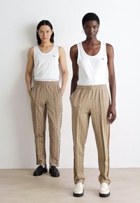 To personer står side om side, iført hvite tank tops og mønstrede brune treningsbukser med sidestriper. Buksene har et geometrisk design.