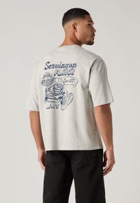 Γκρι βαμβακερό t-shirt με άνετη εφαρμογή, διακοσμημένο στο πίσω μέρος με μπλε καρτούν γραφικό ενός χαμογελαστού χ�αρακτήρα με στοιχεία φαγητού και κείμενο.