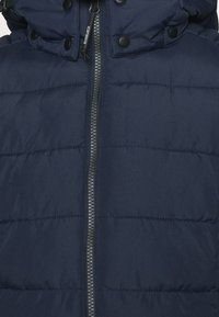 Doudoune marine dotée d'une fermeture éclair intégrale, d'un design matelassé, de boutons-pression noirs et d'un tissu texturé.