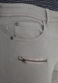 Graue Baumwollhose mit einer Vordertasche, einem metallenen Reißverschluss mit silbernen Zähnen und kontrastierenden Nähdetails. Glatte Textur und maßgeschneiderter Schnitt.
