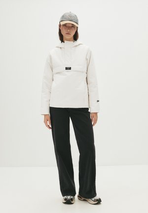 Pullover bianco con cappuccio e tasca frontale, abbinato a pantaloni neri a gamba larga e sneakers con motivo. Un cappellino da baseball grigio completa l'outfit.