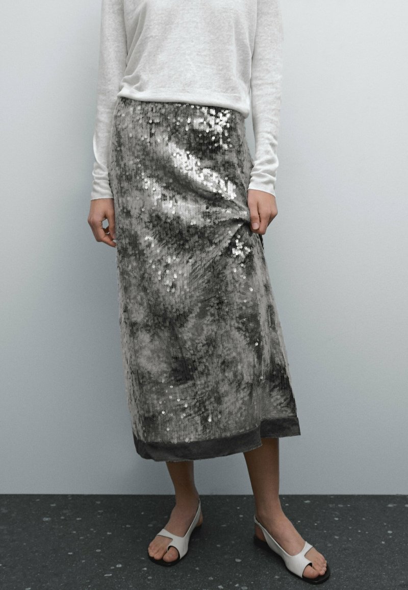 Jupe midi pailletée en tons argentés et gris, présentant une texture lisse et une coupe droite, assortie à un haut blanc à manches longues et des sandales.