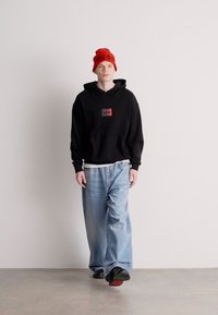 Μαύρη φόρμα με μικρή κόκκινη ετικέτα, συνδυασμένη με φαρδιά μπλε τζιν παντελόνια. Το μοντέλο φοράει ένα φωτεινό κόκκινο σκουφάκι και μαύρα παπούτσια.