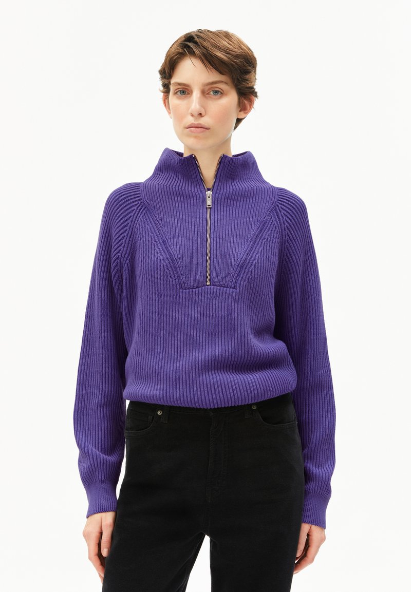 Lila Rippstrickpullover mit hohem Kragen und halbem Reißverschluss. Hat ein strukturiertes Design und lange Ärmel, kombiniert mit schwarzen Hosen.