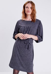 Vestido gris texturizado con una silueta relajada, mangas abullonadas, cuerpo plisado y un detalle de lazo en la cintura. Diseño sencillo hasta la rodilla.