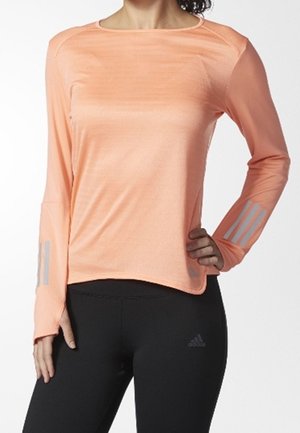 Kobieta ubrana w brzoskwiniową sportową bluzę z długim rękawem i odblaskowymi paskami na rękawach oraz czarne legginsy Adidas.