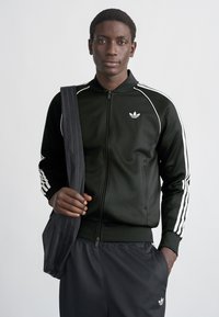 Juoda sportinė striukė su baltomis juostelėmis, užtrauktuku priekyje ir logotipu kairėje krūtinės pusėje, derinama su atitinkančiomis kelnėmis ir tamsia per petį nešiojama rankine.