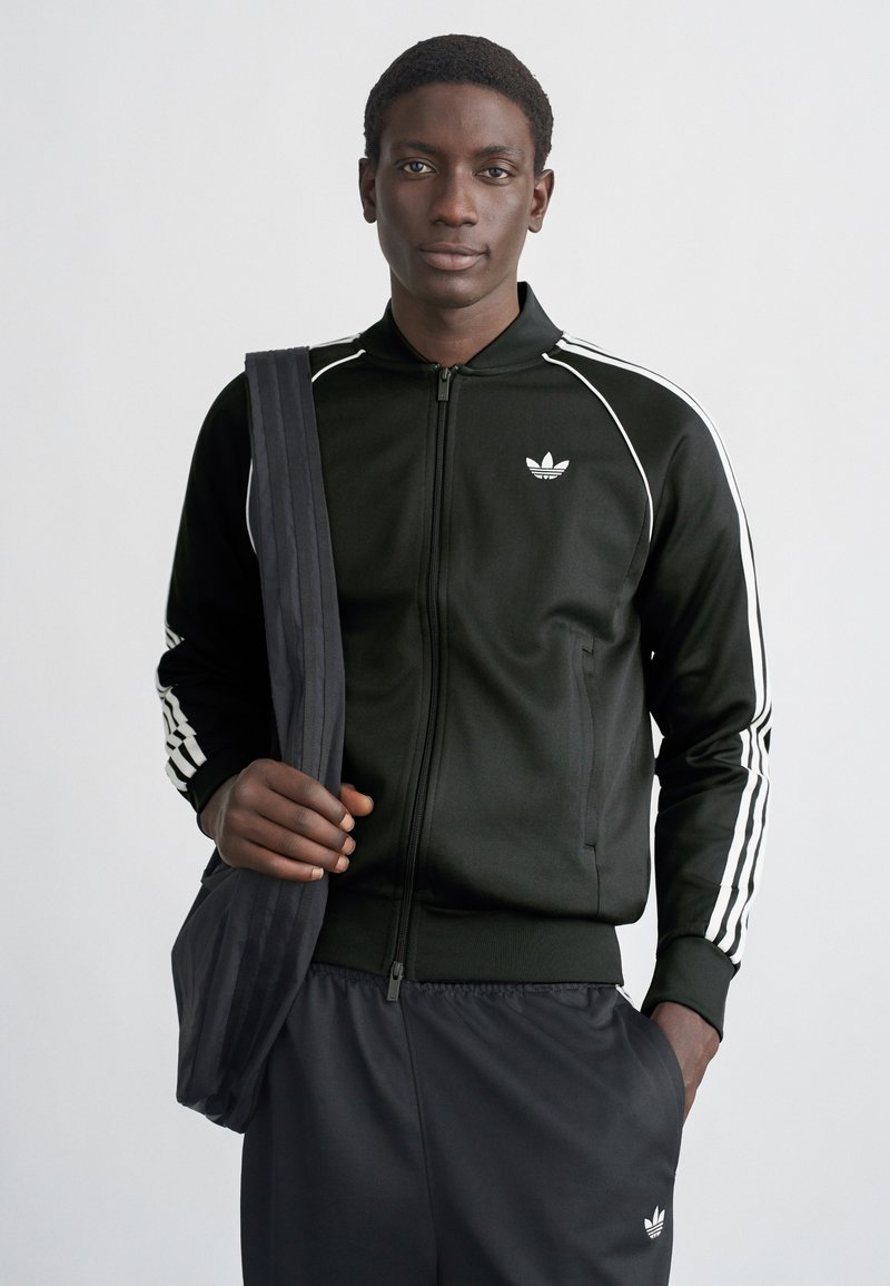 Juoda sportinė striukė su baltomis juostelėmis, užtrauktuku priekyje ir logotipu kairėje krūtinės pusėje, derinama su atitinkančiomis kelnėmis ir tamsia per petį nešiojama rankine.