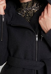 Cappotto in misto lana nero con colletto alto, zip a lunghezza completa e cinturino abbinato. Caratterizzato da una texture liscia e un design elegante.