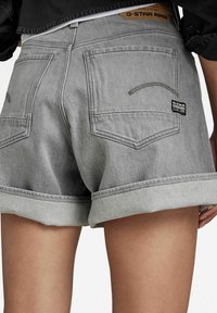 Pantalones cortos de mezclilla gris con dobladillo enrollado. Cuenta con dos bolsillos traseros, una etiqueta de la marca y detalles de costura visibles. Textura suave, diseño casual.