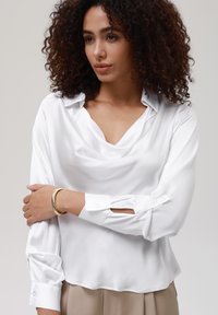 Blusa de satén blanca con escote drapeado, mangas largas con puños abotonados y detalle cortado en el codo. Textura suave.