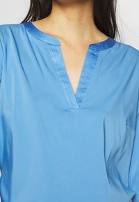 Donna che indossa una blusa azzurro chiaro con un collo a V e dettagli in blu scuro lungo il colletto e il neckline.