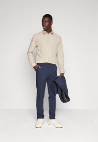 Helles braunes Langarmhemd, marineblaue Hose, hält eine marineblaue Jacke. Weiße Sneakers. Glatter Stoff, taillierte Passform, einfaches Knopfdesign.