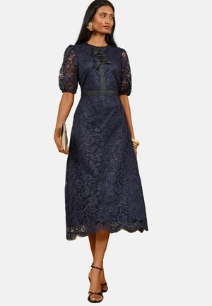 Mujer con un vestido midi de encaje azul marino con mangas abullonadas, un detalle de lazo negro en el pecho, sosteniendo un bolso de mano y con sandalias de tacón alto.
