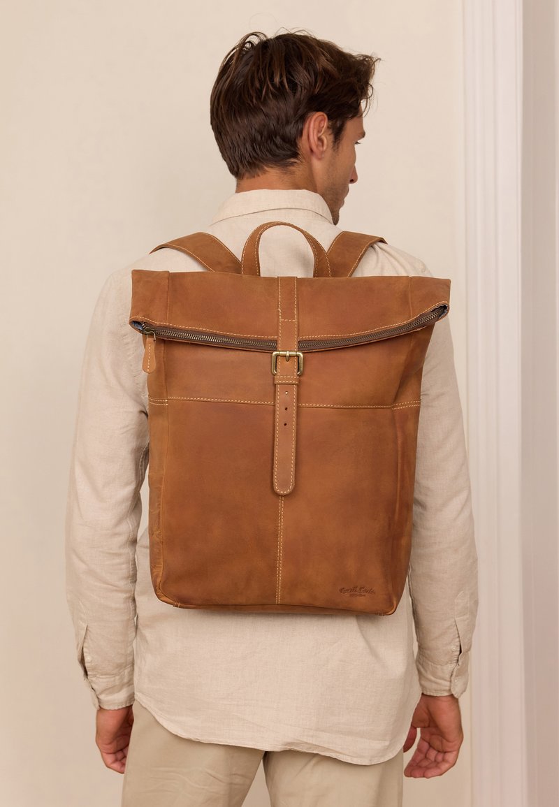 Ruda odinė kuprinė su suvyniojamu viršumi, užtrauktuku ir žalvario sagtimi. Yra siūlių akcentai ir reguliuojamos petnešos.