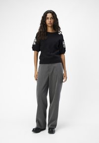 Suéter negro de punto con bordado floral en las mangas; pantalones de pierna ancha grises; modelo con zapatos negros; fondo neutro.