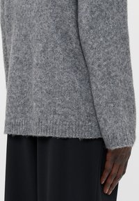 Maglione grigio a maglia con una vestibilità ampia e orlo a costine, caratterizzato da una texture morbida. La vista laterale mostra la cucitura dettagliata e la lunghezza delle maniche.