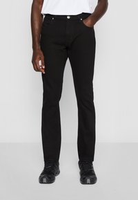Homme portant un jean noir slim, chemise blanche rentrée et baskets noires, debout avec une main dans la poche sur fond gris uni.