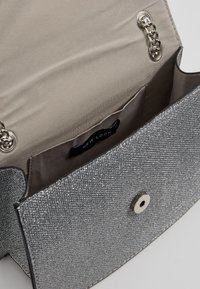 Borsa a tracolla argentata con una finitura testurizzata e un riflesso metallico, dotata di un interno beige, una chiusura a scatto magnetico e un'etichetta del marchio.