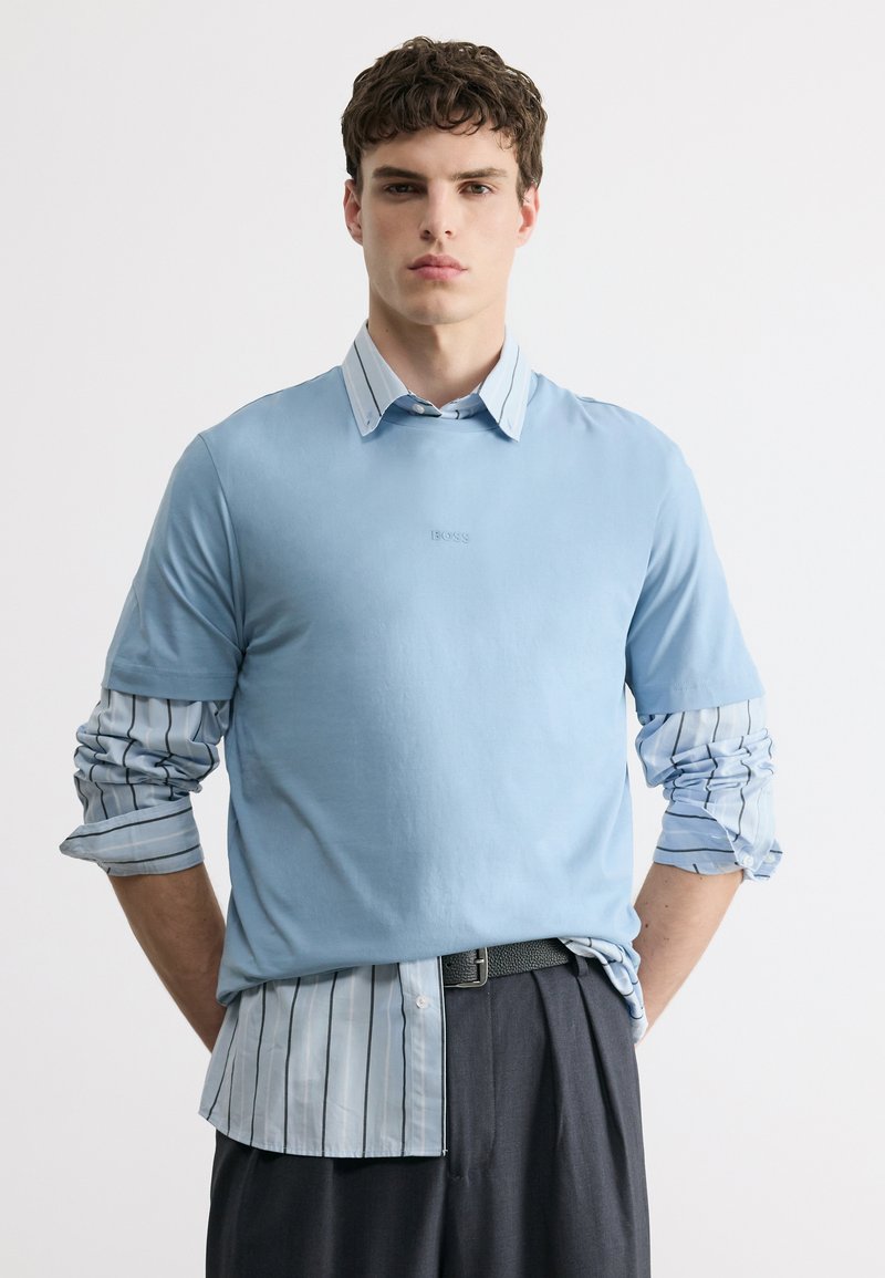 Νέος άνδρας με κοντά σγουρά μαλλιά που φοράει ελαφρύ μπλε κοντομάνικο πουκάμισο πάνω από ένα μπλε και λευκό ριγέ μακρυμάνικο πουκάμισο, όρθιος.