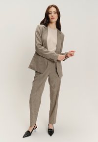 Completo da donna a quadri: motivo houndstooth marrone chiaro e marrone scuro, vestibilità su misura, giacca monopetto, pantaloni con davanti piatto, abbinati a tacchi neri.