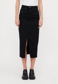 Fekete farmer midi szoknya elülső hasítékkal, magas derékkal, öt zsebes kialakítással és egyenes szabással. Fekete slip-on cipővel párosítva.