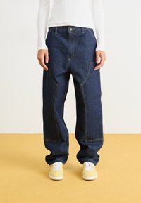 Dark denim trousers with a loose fit, featuring yellow stitching, front pockets, and a high waist design, paired with yellow sneakers.