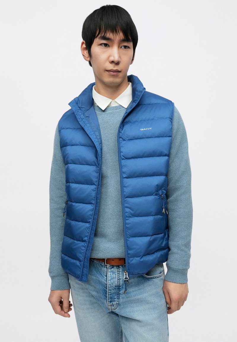 Blue padded vest over a light blue sweater, featuring zippered pockets, a high collar, and a smooth texture with subtle branding.