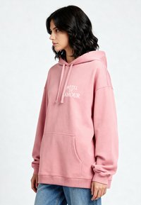 Felpa rosa con tasca frontale e testo bianco "HÔTEL DE L'AMOUR." Maniche ampie e orlo a costine, realizzata in un morbido tessuto testurizzato.