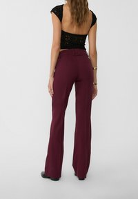 Bordowe spodnie flare z wysokim stanem, wykonane z gładkiego materiału, z pętelkami na pasek i starannie wykończonym dołem. Widok z tyłu.